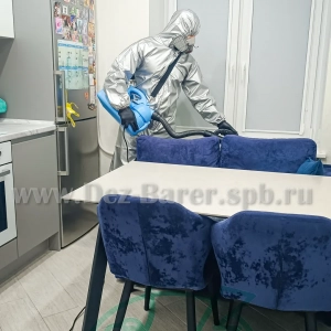 Дезинфектор в защите обрабатывает синие стулья и стол на кухне