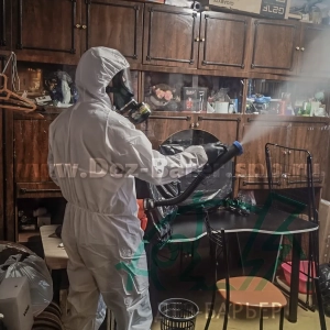 дезинсектор в костюме и маске направляет струю распылителя на мебель у шкафа