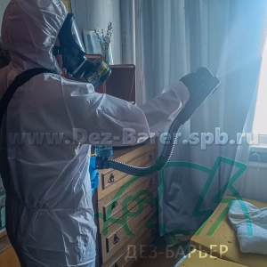 Дезинсектор в защитном костюме и респираторе обрабатывает зону рядом с комодом